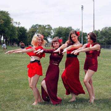 Four femme people in red outfits to resemble Kate Bush. They are in a green field.